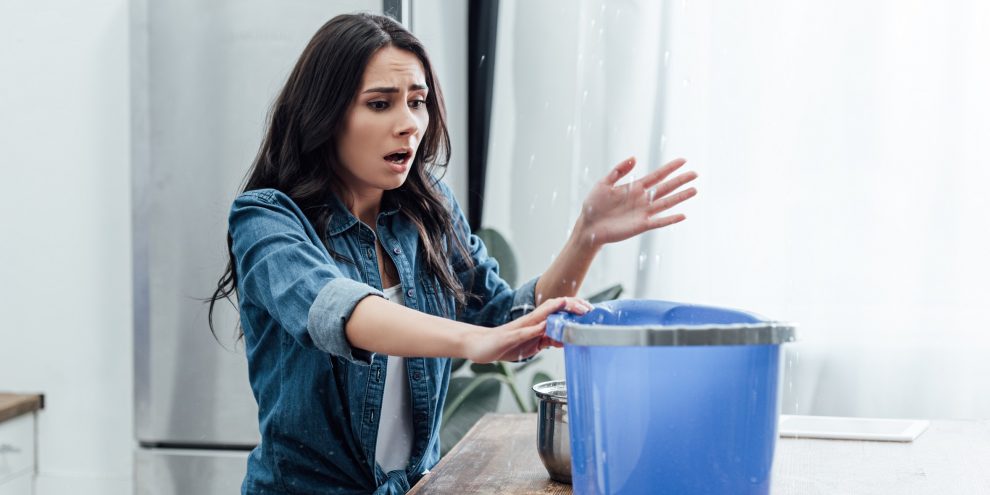 Upset young woman using bucket during leak in kitchen